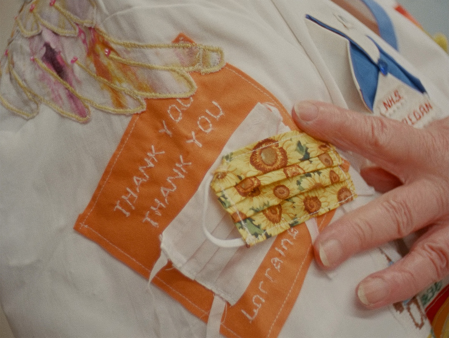 16mm film still from Strathearn by Helen McCrorie, close-up of embroidered nhs scrubs, featuring wings and a face-mask that the wearer points to